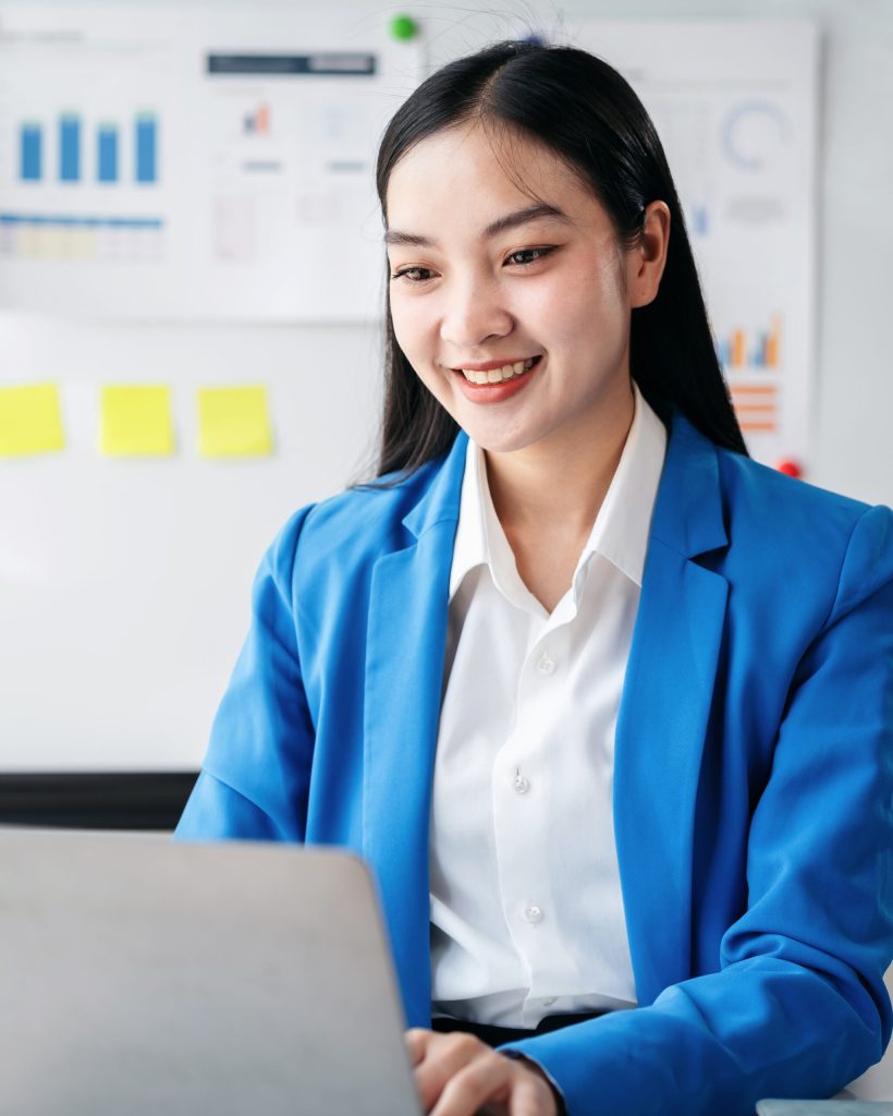 A woman in a blue suit is sitting at a desk with a laptop in front of her. She is smiling and she is happy